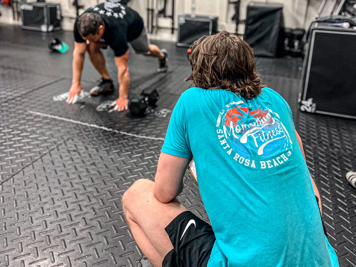 A coach in a Momentum Fitness shirt watching a member&rsquo;s form