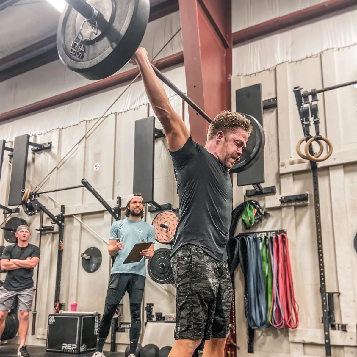 A member locking out a barbell overhead while a judge watches